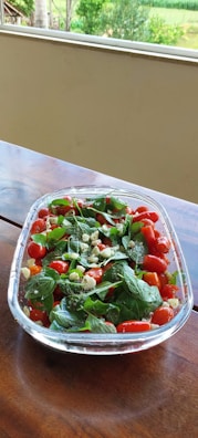 A clear glass bowl filled with a fresh salad consisting of cherry tomatoes, basil leaves, and pieces of cheese sits on a wooden table. The vivid colors of the ingredients stand out against the backdrop of a green garden visible through a large window.