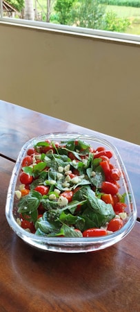 A clear glass bowl filled with a fresh salad consisting of cherry tomatoes, basil leaves, and pieces of cheese sits on a wooden table. The vivid colors of the ingredients stand out against the backdrop of a green garden visible through a large window.