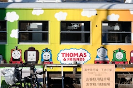 A colorful train carriage features illustrations of cartoon trains with smiling faces. The sides of the carriage are painted in bright shades of yellow and green, with white clouds and vibrant characters from a popular children’s show. A sign in Japanese is visible in the foreground, along with parked bicycles.