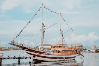 A large wooden sailing ship docked at a pier, adorned with colorful triangular flags along its masts. The ship is reflected in calm water, and there are buildings and palm trees visible in the background under a partly cloudy sky.