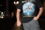 A person stands in a dimly lit urban environment wearing a black Iron Maiden T-shirt featuring the 'Seventh Son of a Seventh Son' album art. The background shows blurred lights and architectural details, suggesting it is night time.