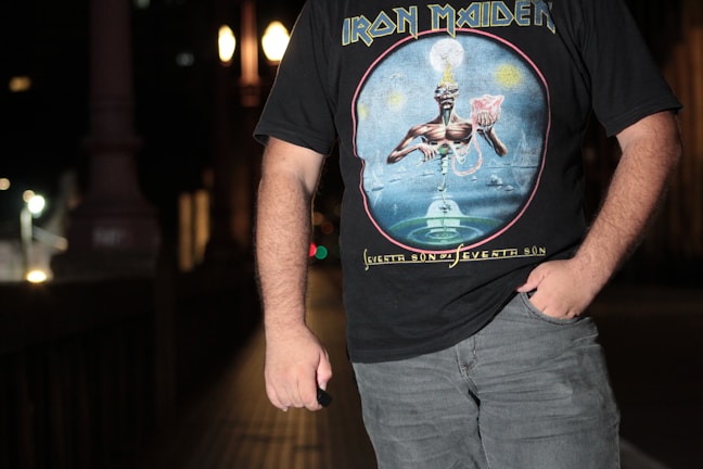 A person stands in a dimly lit urban environment wearing a black Iron Maiden T-shirt featuring the 'Seventh Son of a Seventh Son' album art. The background shows blurred lights and architectural details, suggesting it is night time.