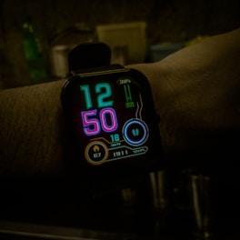A digital smartwatch displays the time 12:50 with futuristic neon-colored numbers on a black background. Various health and activity metrics are visible below the time, including date, heart rate, and steps taken, indicated by glowing icons and numbers.