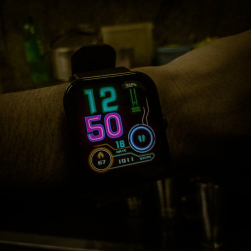 A digital smartwatch displays the time 12:50 with futuristic neon-colored numbers on a black background. Various health and activity metrics are visible below the time, including date, heart rate, and steps taken, indicated by glowing icons and numbers.