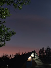 A peaceful evening scene featuring a triangular-roofed building illuminated by a soft light. The sky transitions from a pinkish hue near the horizon to a deeper blue further up. Silhouettes of trees frame the scene, and the branches of a tree with green leaves are visible.