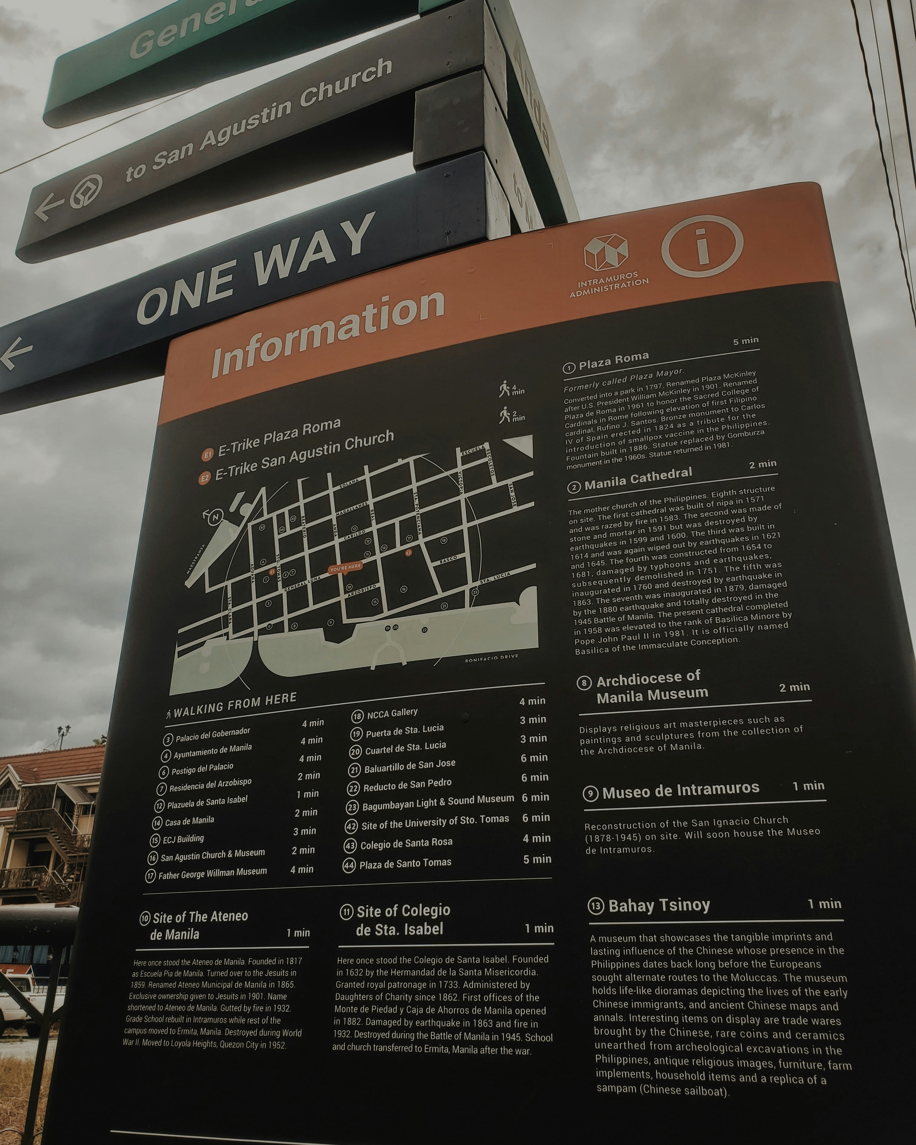 A tall information board with a map and detailed descriptions of various locations, including historical sites and museums, situated in an outdoor area. The board is positioned under street signs that guide towards San Agustin Church and indicate one-way traffic. The sky appears overcast, and part of a building is visible in the background.