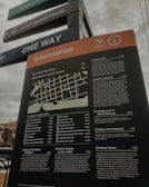 A tall information board with a map and detailed descriptions of various locations, including historical sites and museums, situated in an outdoor area. The board is positioned under street signs that guide towards San Agustin Church and indicate one-way traffic. The sky appears overcast, and part of a building is visible in the background.