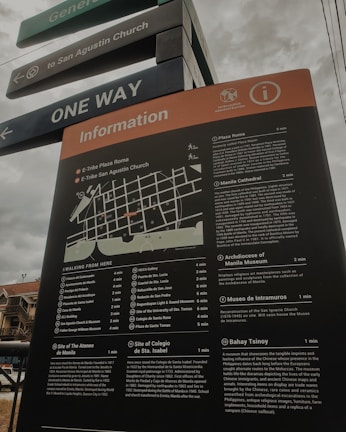 A tall information board with a map and detailed descriptions of various locations, including historical sites and museums, situated in an outdoor area. The board is positioned under street signs that guide towards San Agustin Church and indicate one-way traffic. The sky appears overcast, and part of a building is visible in the background.