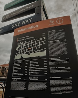 A tall information board with a map and detailed descriptions of various locations, including historical sites and museums, situated in an outdoor area. The board is positioned under street signs that guide towards San Agustin Church and indicate one-way traffic. The sky appears overcast, and part of a building is visible in the background.