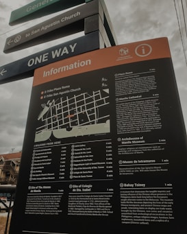 A tall information board with a map and detailed descriptions of various locations, including historical sites and museums, situated in an outdoor area. The board is positioned under street signs that guide towards San Agustin Church and indicate one-way traffic. The sky appears overcast, and part of a building is visible in the background.