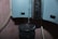 A close-up of a water dispenser with a droplet of water falling from the spout into a dark blue cup. The background has a light pink floral patterned wallpaper. The dispenser appears metallic and reflective.