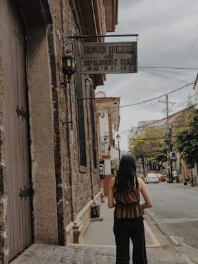 A person with long hair stands on a cobblestone street next to an old building with textured brick walls. A sign hangs from the building reading 'Ironcon Builders and Development Corp.' Overhead power lines and trees line the street, which extends into the distance with a few parked cars visible.