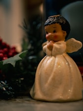 A small ceramic angel figurine with black curly hair and rosy cheeks, posed with hands clasped in prayer. The figure is wearing a cream-colored, sparkly gown with wings. It is surrounded by festive, green foliage and some red holly berries in a softly focused background.