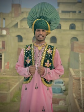 A person is wearing traditional attire with a vibrant headpiece and intricate embroidery on the vest and tunic. The background features an unfinished or rustic building with visible bricks and a car parked behind. The overall setting gives off a cultural and festive vibe.