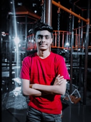 A young person wearing a red shirt stands confidently with arms crossed in front of a playground structure. The background is dimly lit with bright spots of light creating dramatic contrasts. The playground equipment is mostly dark with some orange elements.