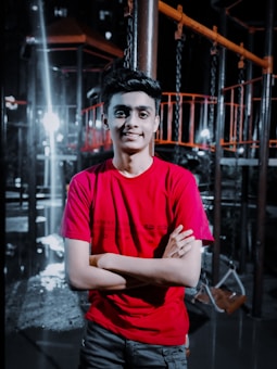 A young person wearing a red shirt stands confidently with arms crossed in front of a playground structure. The background is dimly lit with bright spots of light creating dramatic contrasts. The playground equipment is mostly dark with some orange elements.