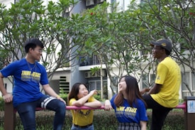 Four people are gathered outdoors, appearing to enjoy each other's company. Two of them are wearing blue T-shirts, while the other two are wearing yellow T-shirts, all with similar logos. The group is positioned under green trees, and they are smiling and engaging in conversation. The background includes a building with windows.