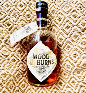 A bottle of Woodburns Contemporary Indian Whisky with a label featuring intricate design elements and text. The bottle cap is labeled with 'GOA INDIA'. The background consists of a textured pattern in beige and white.