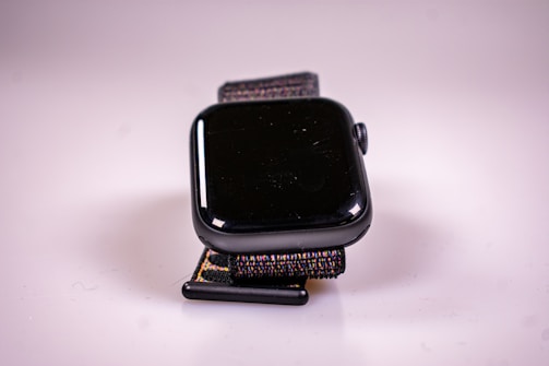 A close-up view of a black smartwatch with a reflective screen and a woven fabric strap. The watch is positioned upright against a simple, light-colored background.