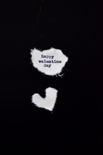 Two pieces of white fabric are arranged on a black background. The upper piece is irregularly shaped and contains the text 'happy valentine day' in dark blue. Below it, there is a smaller fabric piece shaped like a heart.