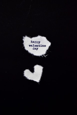 Two pieces of white fabric are arranged on a black background. The upper piece is irregularly shaped and contains the text 'happy valentine day' in dark blue. Below it, there is a smaller fabric piece shaped like a heart.