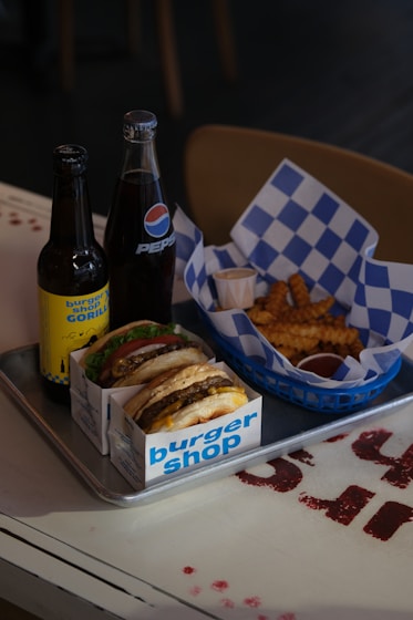 A tray containing two burgers in branded packaging, a serving of crinkle-cut fries in a basket with a blue and white checkered paper liner, and two bottles of soda: one bottle of Pepsi and another with a custom label. There are also two small cups of dipping sauce next to the fries.