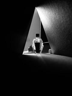 The image is a black and white photograph featuring a geometric composition. A small bottle with an angular design is positioned at the center, framed by two dark, triangular shapes converging at the top. Soft light is illuminating the bottle, creating contrast against the shadows and highlighting its distinctive shape.