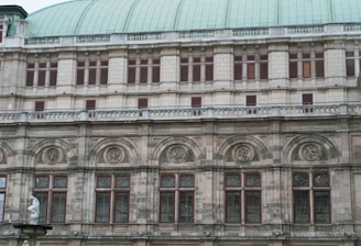 The image features a section of a historic building facade with intricate architectural details. The structure showcases a series of arched windows with decorative stonework and carvings above them. A portion of a statue is visible at the bottom left corner, adding to the classical aesthetic.