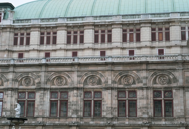 The image features a section of a historic building facade with intricate architectural details. The structure showcases a series of arched windows with decorative stonework and carvings above them. A portion of a statue is visible at the bottom left corner, adding to the classical aesthetic.