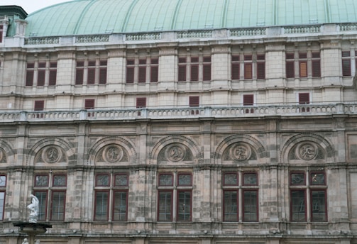The image features a section of a historic building facade with intricate architectural details. The structure showcases a series of arched windows with decorative stonework and carvings above them. A portion of a statue is visible at the bottom left corner, adding to the classical aesthetic.