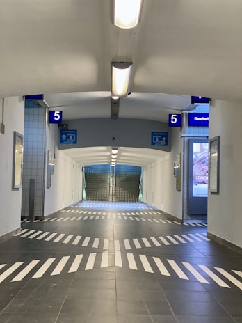 A tunnel-like passageway with overhead fluorescent lighting and dark tiled flooring featuring white arrow markings. Blue signs display the number 5, and there are advertisements on the walls.