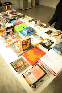 A table filled with an assortment of books, some lying flat while others are stacked. The books cover various topics, including design, art, and biographies. One person's hand is visible, flipping through one of the books. The background includes a minimalist room with concrete flooring.