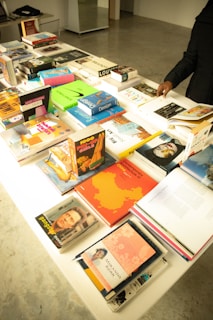 A table filled with an assortment of books, some lying flat while others are stacked. The books cover various topics, including design, art, and biographies. One person's hand is visible, flipping through one of the books. The background includes a minimalist room with concrete flooring.