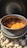 A black pot filled with a rich, orange stew containing chunks of meat and vegetables. The pot is sitting on a bed of light-colored stones, and steam is visible rising from the stew. The lid of the pot is open, with the interior showing signs of cooking.