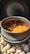 A black pot filled with a rich, orange stew containing chunks of meat and vegetables. The pot is sitting on a bed of light-colored stones, and steam is visible rising from the stew. The lid of the pot is open, with the interior showing signs of cooking.