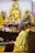 Two monks are seated in a Buddhist temple adorned with golden statues and intricate designs. The focus is on the spiritual atmosphere, with flowers and offerings around. The monks wear traditional yellow and maroon robes.