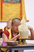 A person wearing a maroon and yellow robe is carefully crafting a large, intricate sculpture on a pedestal. The sculpture appears to be made of a soft material and includes several rounded shapes and protrusions. In the background, a colorful, ornate fabric hangs on the wall. A table in the foreground holds bowls and utensils, suggesting a focus on detailed handiwork.