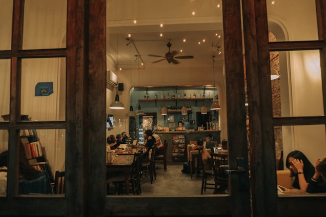 A cozy coffee shop interior viewed through wooden-framed glass doors. The space is warmly lit with overhead lamps and decorated with shelves filled with bottles and books. Several people are seated at tables, engaged in conversation or working on laptops. The walls have a mix of modern and rustic elements, creating an inviting atmosphere.