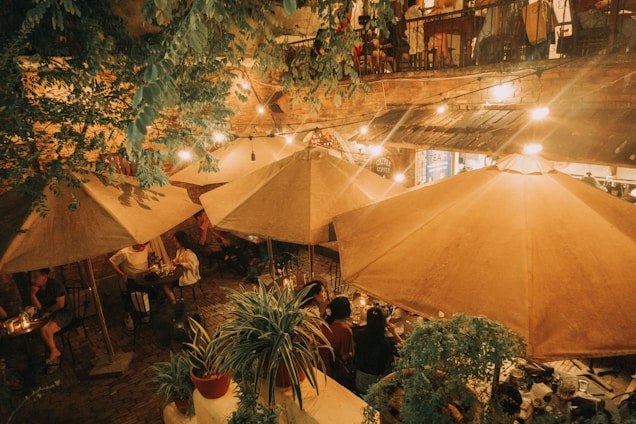 A cozy outdoor café setting with warmly lit string lights and large wooden umbrellas shading tables. Patrons relax at small tables, enjoying food and drinks under the ambient glow framed by greenery. Upstairs features a balcony area with more people gathered, contributing to the bustling, yet intimate atmosphere.