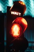 A traffic light displays a red glowing hand and the word 'WAIT' in illuminated letters. The surrounding environment is dimly lit, creating a strong contrast between the light's red hues and the dark background. The image evokes an urban nighttime setting.