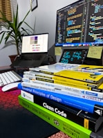 A workspace featuring a computer monitor displaying code, an open laptop showing a design interface, and a stack of books related to programming and design. The environment includes a keyboard, a mouse, sticky notes, and a potted plant, creating a tech-savvy and organized atmosphere.