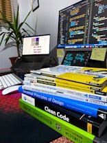A workspace featuring a computer monitor displaying code, an open laptop showing a design interface, and a stack of books related to programming and design. The environment includes a keyboard, a mouse, sticky notes, and a potted plant, creating a tech-savvy and organized atmosphere.