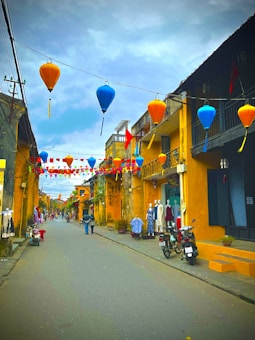 A picturesque street lined with colorful lanterns hangs across the sky. The street is flanked by old, yellow-walled buildings with balconies. Several mannequins display clothing outside shops, and motorcycles are parked nearby. The atmosphere is calm, with a few people strolling down the street.