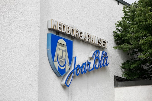 The image features a sign on a building with white textured walls. The sign includes a shield emblem with a stylized portrait of a bearded figure wearing headwear, against a blue background. Above the emblem, the word 'MEDBORGARHUSET' is displayed in uppercase beige letters, and beneath it, the script 'Joar Blå' is written in flowing blue cursive letters. Green leaves from a tree partially cover the right side of the building.