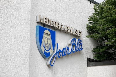 The image features a sign on a building with white textured walls. The sign includes a shield emblem with a stylized portrait of a bearded figure wearing headwear, against a blue background. Above the emblem, the word 'MEDBORGARHUSET' is displayed in uppercase beige letters, and beneath it, the script 'Joar Bl&aring;' is written in flowing blue cursive letters. Green leaves from a tree partially cover the right side of the building.
