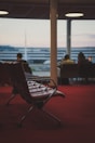 A row of empty airport lounge chairs is positioned on a red carpet. In the background, a large window reveals out-of-focus terminal buildings and people seated, some looking out of the window.