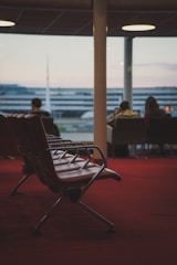 A row of empty airport lounge chairs is positioned on a red carpet. In the background, a large window reveals out-of-focus terminal buildings and people seated, some looking out of the window.