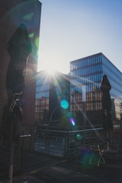 Modern office buildings are illuminated by the sun, casting lens flares across the image. A couple of closed patio umbrellas stand in the foreground on a wooden terrace, while reflections are visible on the glass facades.