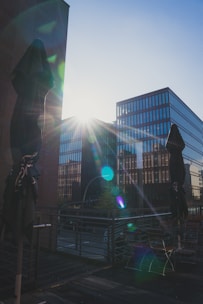 Modern office buildings are illuminated by the sun, casting lens flares across the image. A couple of closed patio umbrellas stand in the foreground on a wooden terrace, while reflections are visible on the glass facades.