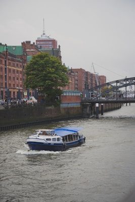 A small blue and white boat with a blue canopy is moving along a river in an urban area. The backdrop features historic brick buildings with green rooftops, a large tree, and a bridge crossing over the river. There are people walking along the riverside, and a tall building with an antenna is visible in the distance.